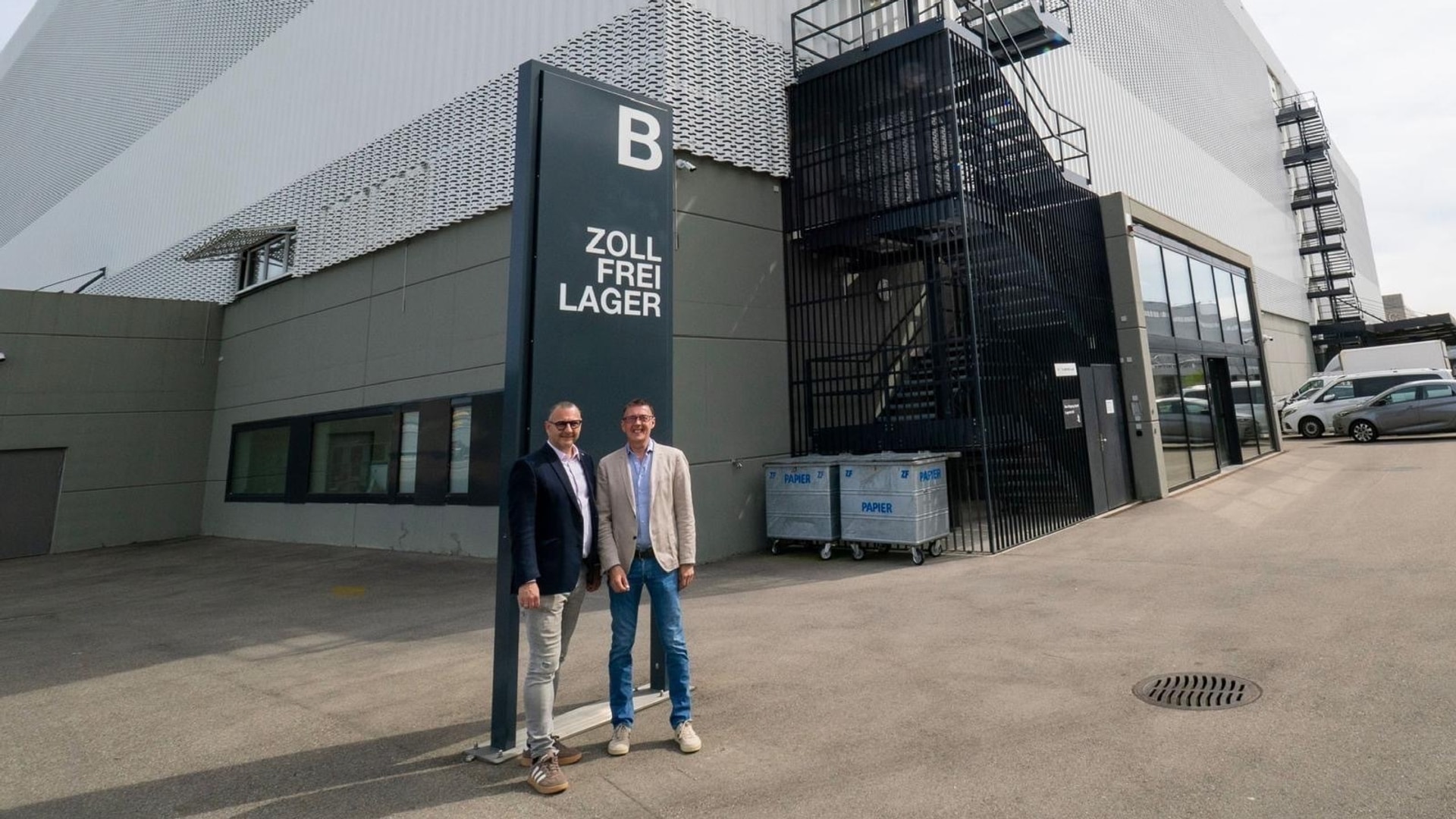Ing. Paul Eichberger und Ing. Christian Weinberger-Prammer vor dem Eingang des Hochsicherheitslagers in der Schweiz, neben einem dunklen Pfeiler mit großem „B“; rechts eine schwarze Außentreppe hinter einem Gitter und ein verglaster Eingang.