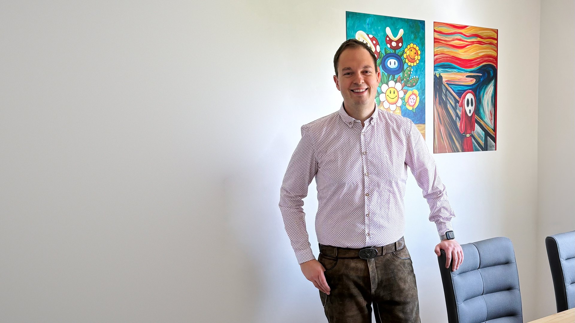 Mag. Stefan Gündhör steht in der rechten Bildhälfte vor zwei Gemälden, in hellem Hemd und Lederhose, eine Hand auf der Lehne eines Stuhls; die linke Bildhälfte ist helle, freie Wand.